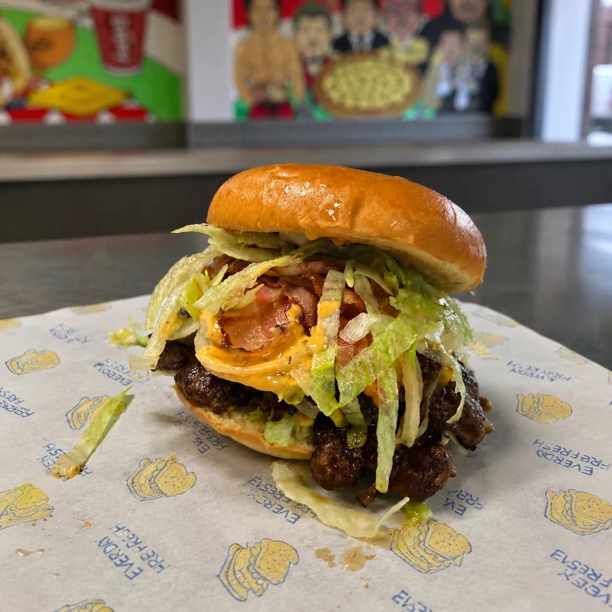 Loaded bacon cheeseburger with crispy bacon, shredded lettuce, melted cheese, and creamy sauce in a glossy brioche bun, served on branded burger paper with a softly blurred takeaway restaurant background.